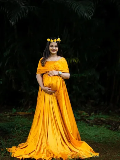 A full-length portrait of a mom-to-be in a golden-yellow gown, standing confidently in a beautiful, dark, and leafy setting.