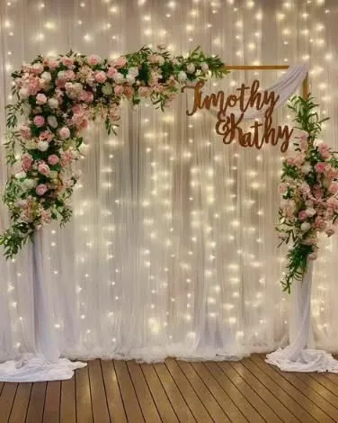 A simple and elegant photobooth with a backdrop of white curtains and fairy lights. The asymmetrical floral arrangement and custom couple's name sign add a personal and romantic touch.