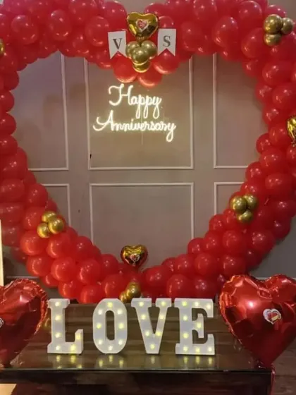 I design grand romantic gestures for anniversaries. This setup features a massive heart-shaped arch made of red balloons, a 'Happy Anniversary' neon sign, and illuminated marquee letters spelling 'LOVE' for an unforgettable surprise.