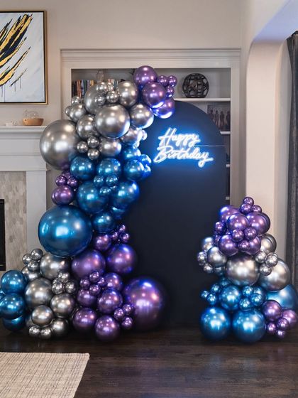 A stunningly modern birthday setup with an organic balloon arch in metallic silver, blue, and purple against a sleek black backdrop with a neon sign.