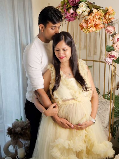 This candid moment shows the couple sharing a quiet connection. He gently kisses her forehead as they both cradle the bump, set against the warm, floral-adorned boho backdrop.