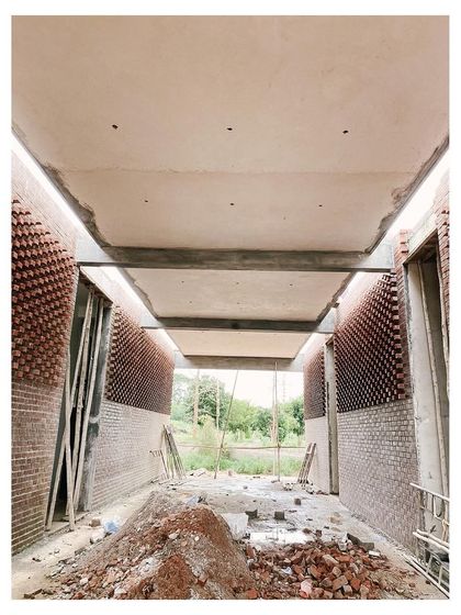 The interior of a hallway during construction, showing the contrast between the smooth concrete ceiling and the textured brick walls. The open end of the corridor frames a view of the natural landscape outside.