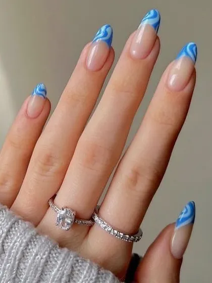 This trendy manicure features a blue wave or swirl pattern on the tips of long, almond-shaped nails. It's a modern and eye-catching take on the classic French tip design.