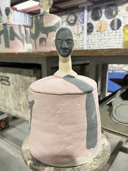 A 'Human Jar' before the final glaze firing. The piece combines a sculpted head as the lid with a pink and grey painted body, showcasing a blend of sculpture and painting.