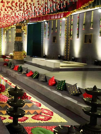 Another angle of the tiered seating, showing the stone lamps and the floral rangoli on the floor. The walls feature a green textured pattern, and floral garlands hang from the ceiling, adding layers of detail to the space.