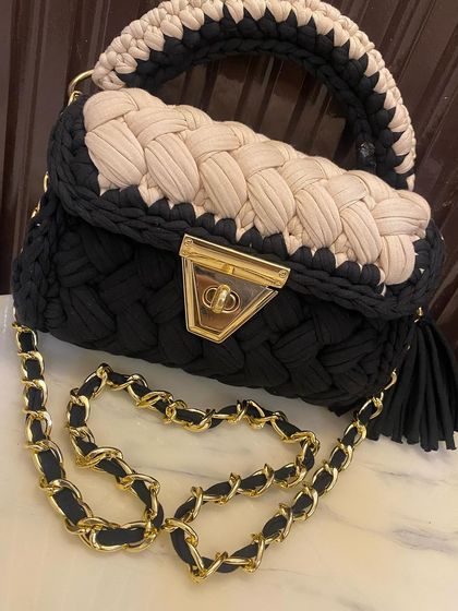 A close-up of the black and beige crochet bag, highlighting the braided texture and the secure gold-tone clasp. The tassel is also handmade with the same yarn.