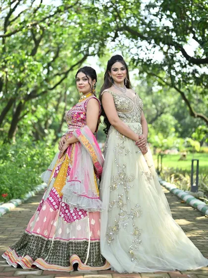 A back-to-back pose featuring a colorful patchwork lehenga and a sophisticated embroidered gown, perfect for sisters or friends attending a wedding.