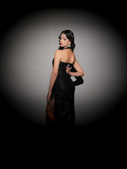 A mysterious and cinematic shot. The model is framed within a circular spotlight, her back to the camera, creating intrigue and highlighting the elegant shape of the black gown.