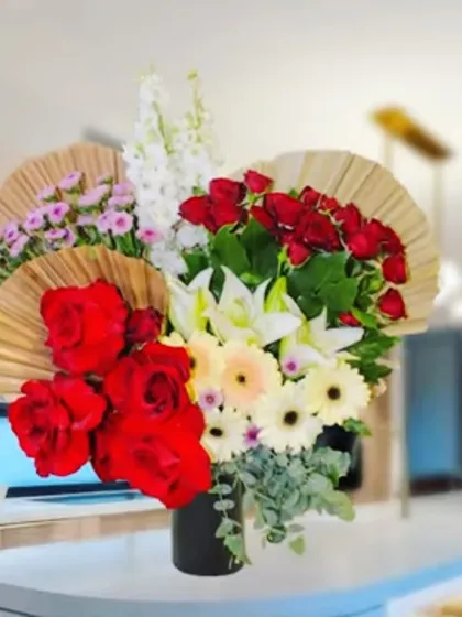 An elegant arrangement of red roses, white lilies, and daisies in a black vase, accented with dried fans. This style is perfect for modern home or office decor.