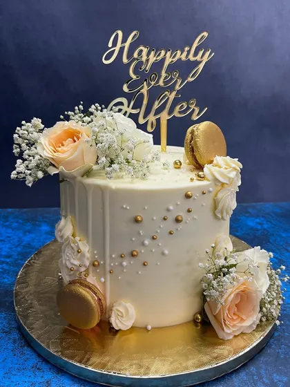 A classic white drip cake for a 'Happily Ever After' moment. This elegant design is adorned with fresh peach roses, baby's breath, and shimmering gold macarons, perfect for engagements, anniversaries, or intimate weddings.