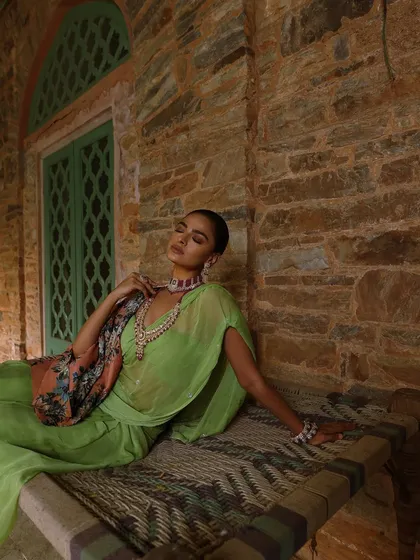 A relaxed, reclining pose that showcases the beautiful drape of the saree and the intricate details of the jewellery against a rustic stone wall.