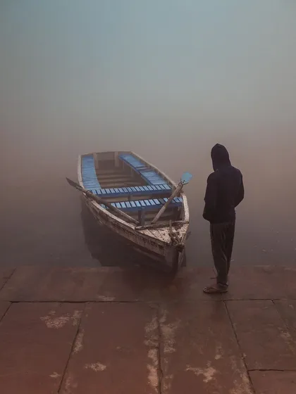 A person in a hoodie stands by a boat on the misty Yamuna ghat, a quiet, introspective moment captured in the soft, diffused light of dawn.