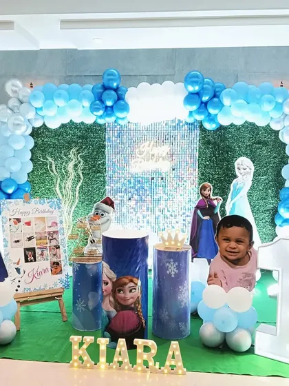 A Frozen-themed first birthday party with a grass-like backdrop and a silver sequin panel. The setup includes character cutouts and a milestone photo board.