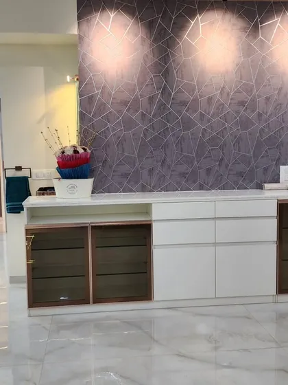 A custom-built crockery and storage unit with a textured wallpaper backdrop. The unit features white lacquer cabinets and smoked glass doors with rose gold frames.