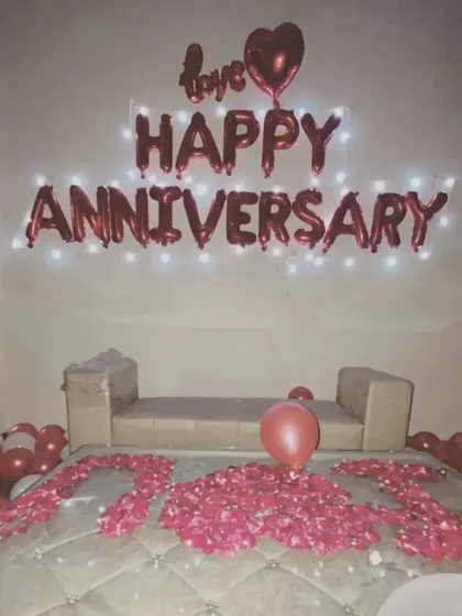 A simple yet heartfelt anniversary decoration. The wall features "Happy Anniversary" foil balloons with lights, and the bed is decorated with rose petals.