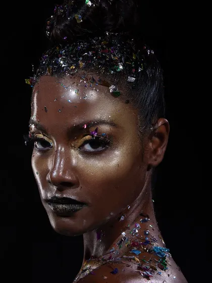 A creative look featuring a deep skin tone, metallic makeup, and scattered glitter accents on the hair and body. We celebrate diversity and creativity in all our shoots.