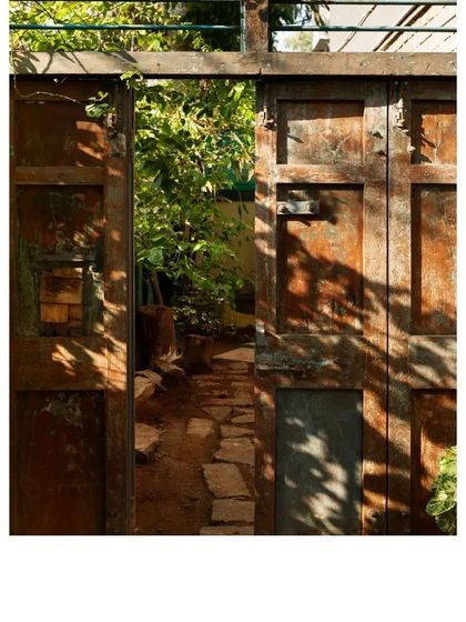 A weathered, rustic door opens to a garden path. The dappled sunlight highlights the texture and history of the upcycled wood, creating a seamless transition between the built and natural worlds.