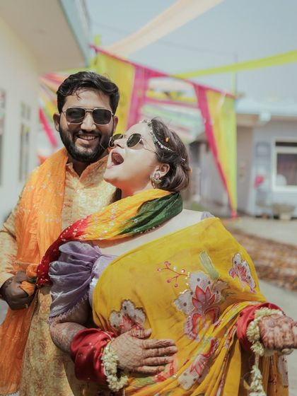 A candid shot of a couple laughing and having fun outdoors during their haldi or mehendi function, showcasing their vibrant outfits and joyful interaction.