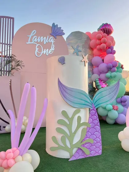 A close-up of the outdoor mermaid party decor, showing a large mermaid tail prop and seashell cutouts against a pastel pink backdrop.