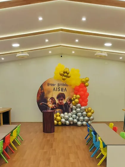 A Harry Potter themed birthday party setup in a party hall, featuring a large circular backdrop with characters from the movie and balloons in house colors.