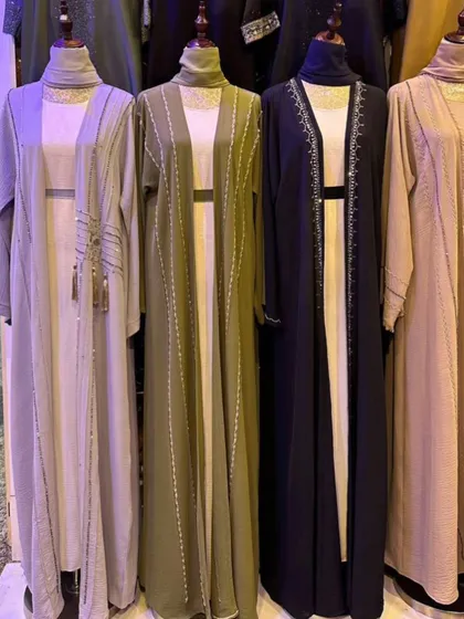 A collection of open front abayas in neutral tones like beige, olive green, and navy. These versatile pieces can be layered easily and feature subtle vertical beading and tassel details.