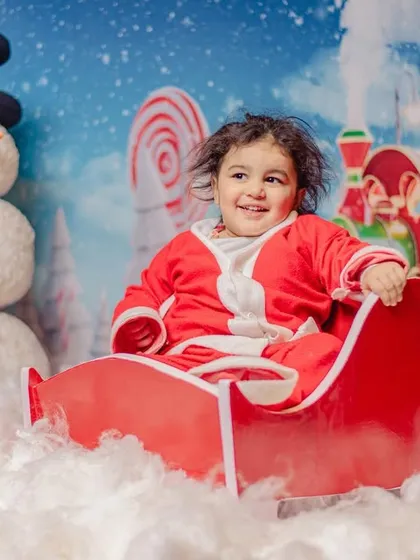 A sweet, smiling portrait of the little Santa in his sleigh, set against a winter wonderland backdrop with a snowman and Christmas tree.
