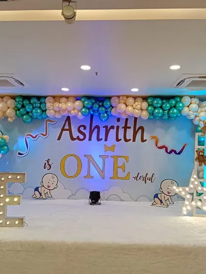An "Ashrith is ONEderful" theme for a first birthday party. The stage is decorated with a large backdrop, marquee letters spelling "ONE," and cute baby illustrations.