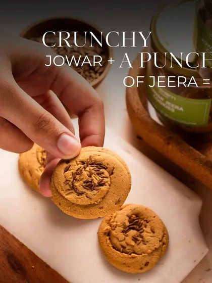 A hand reaches for a Jowar Jeera cookie, showcasing its perfect, crumbly texture. These cookies are my take on a traditional favourite, made with wholesome jowar flour and a punch of jeera for that perfect chai-time snack.