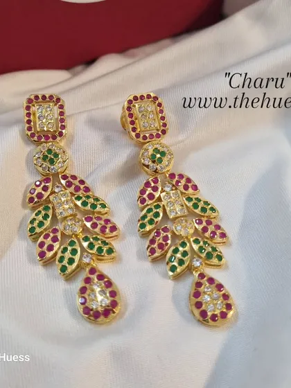 A clear product shot of the 'चारु' (Charu) earrings, showcasing the ruby and emerald coloured stones in a cascading leaf pattern.