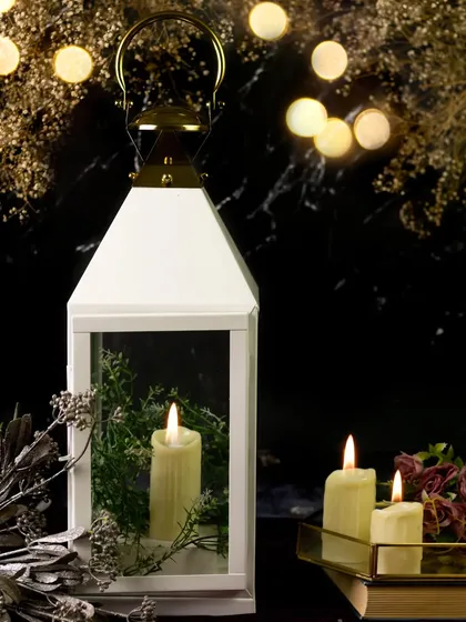 A white lantern is set against a dark background with festive bokeh lights. The addition of greenery inside the lantern and surrounding candles creates a magical, holiday-inspired scene.