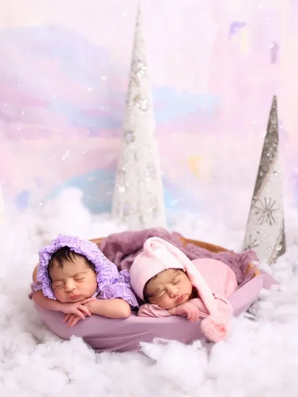 Twin newborns, dressed in adorable purple and pink sleepers, are captured sleeping side-by-side in a winter wonderland setup. This photo beautifully showcases my experience in handling and posing multiple babies for a magical twin photoshoot.