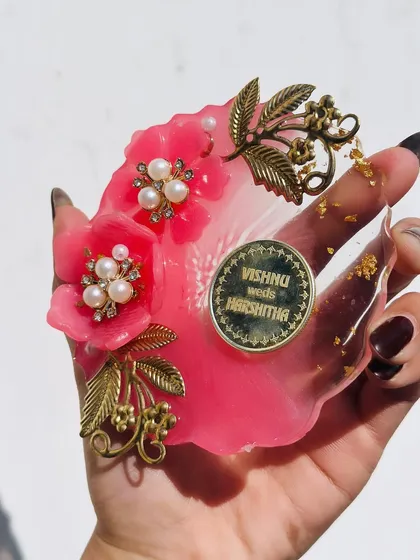 This is a close up of one of my Milni coin plates. I design these decorative resin plates especially for the Milni ceremony at weddings. This one is a vibrant pink flower shape, adorned with pearls, gold-leaf charms, and a custom coin with the couple's names.