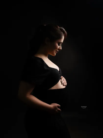 A soft and gentle low-key portrait. The light delicately illuminates the client's smiling profile and her hands on her bump, creating a warm and intimate fine art image.