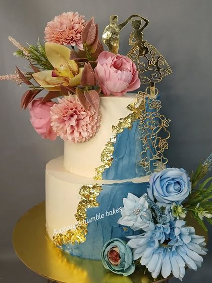 A two-tiered wedding cake with a blue watercolor and gold leaf design, topped with a beautiful arrangement of flowers.