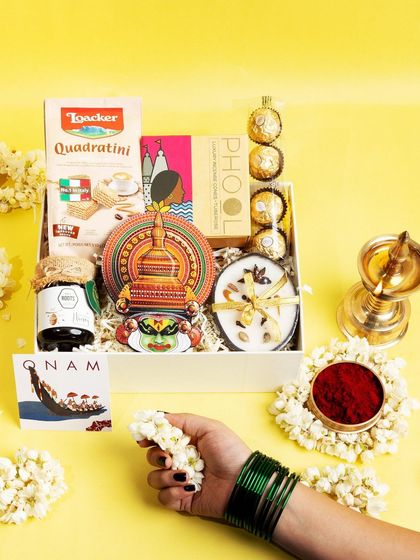 A hand adorned with green bangles reaches towards the 'Jamanthi Poo' hamper, adding a human touch and evoking the festive spirit of Onam.