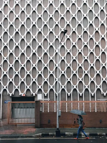 A person with an umbrella walks past a building with a striking geometric facade. The contrast between the organic human figure and the rigid, repeating pattern of the wall makes for a compelling minimalist shot.