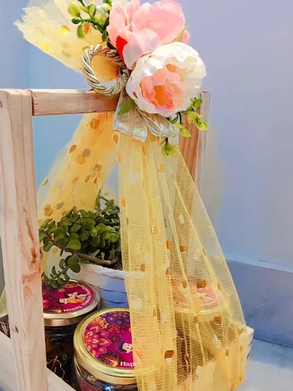 A beautiful Rakhi hamper in a wooden crate, containing jars of treats and a small plant, wrapped in yellow net.