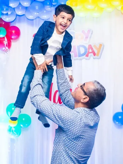 A grandfather joyfully lifts his grandson in the air against a backdrop of colorful birthday balloons.