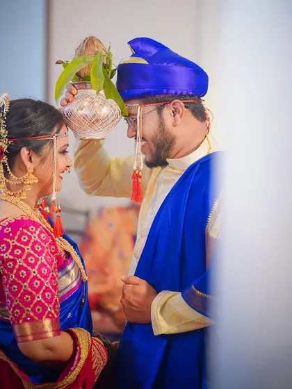 A beautiful moment from a Maharashtrian wedding vidhi. The groom holds a sacred kalash over the bride's head, and their shared smile captures the happiness of the ritual.