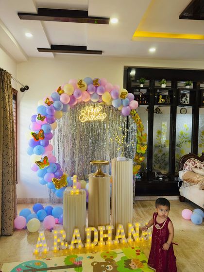 A pastel butterfly theme for a first birthday, with the birthday girl posing next to the decor. The setup includes a circular frame with a silver shimmer backdrop, a multi-color balloon arch, and light-up letters.