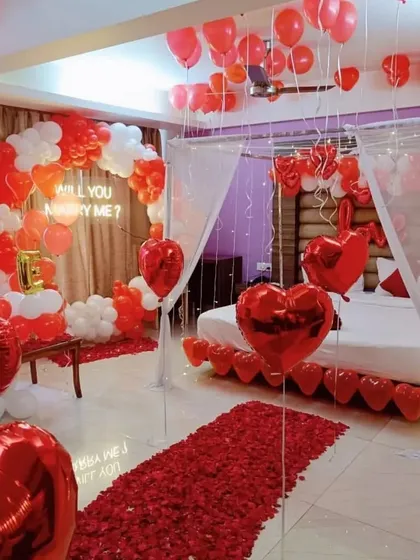A romantic marriage proposal room decoration designed for a truly special moment. The setup features a "Will you marry me?" neon sign, a balloon arch in red and white, a rose petal walkway, and a beautifully decorated bed under a canopy of fairy lights.