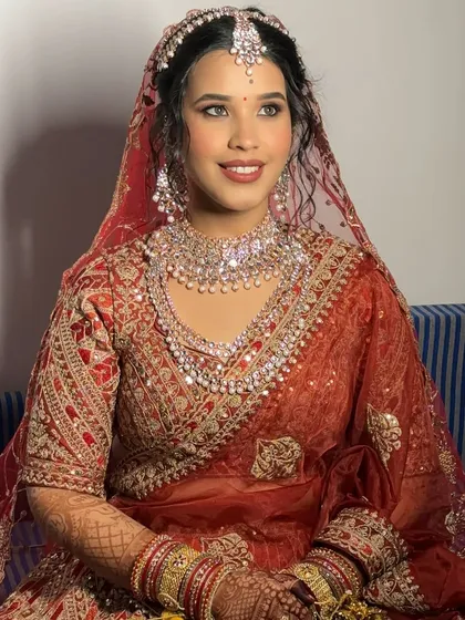 A beautiful seated portrait of the bride in her red lehenga. The makeup features classic elements like defined eyes and a bold lip, perfect for a traditional ceremony.