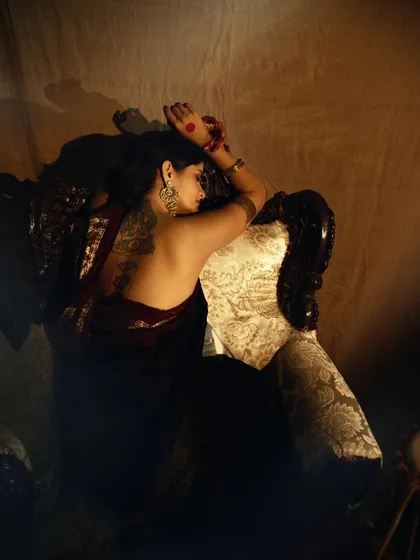 A back-facing portrait that reveals a large, intricate tattoo, blending modern body art with the traditional aesthetic of the saree and setting.