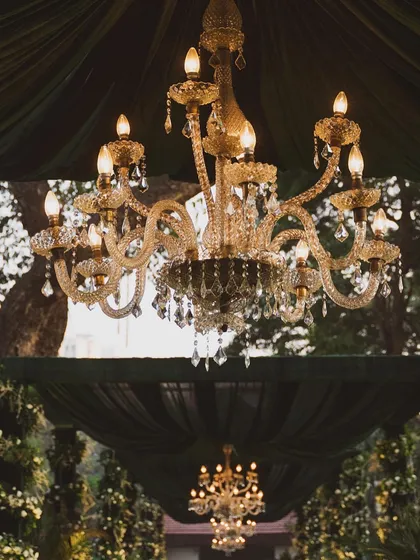 A close-up of a crystal chandelier hanging amidst dark green velvet drapes. This detail shot captures the moody, romantic, and luxurious atmosphere of an enchanted garden-themed wedding.