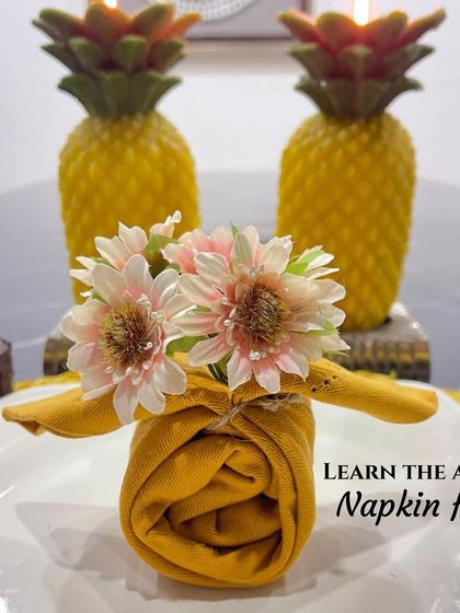 Learn the art of napkin folds with this sunny, pineapple-themed tablescape. The yellow napkin is folded into a rose and topped with a flower, perfectly complementing the pineapple-shaped candles in the background.
