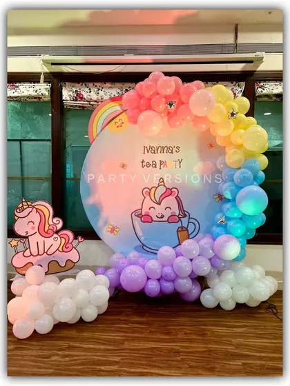 A whimsical Unicorn tea party theme. The centerpiece is a custom circular backdrop with a rainbow balloon garland, complemented by cute unicorn cutouts.