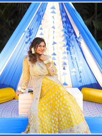 The bride looking beautiful in her yellow Haldi outfit, posing against a vibrant blue and white themed decor.