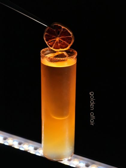 Our Golden Affair cocktail is a tall, elegant drink with a warm glow. It features a complex blend of flavors, finished with a Tajin-dusted rim and a dehydrated orange slice for a touch of spice and citrus aroma.
