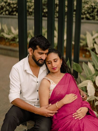 A beautiful outdoor maternity portrait of a couple in a garden setting. The woman's traditional pink saree adds a vibrant touch of color to the natural surroundings.