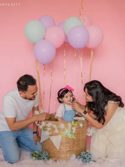 This first birthday shoot uses a lovely pastel palette with a pink background and soft-colored balloons. The parents lovingly guide their daughter in the hot air balloon basket.